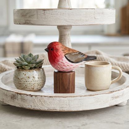 A decorative wooden Purple finch figurine with a red, pink, and brown body perched on a small dark wood block, styled on a distressed white two-tier tray alongside a small potted succulent and a ceramic mug.