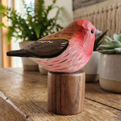 A handcrafted wooden Purple finch figurine with a pink and brown painted body, perched on a round wooden base and displayed on a rustic wooden table with potted plants in a softly lit indoor setting.