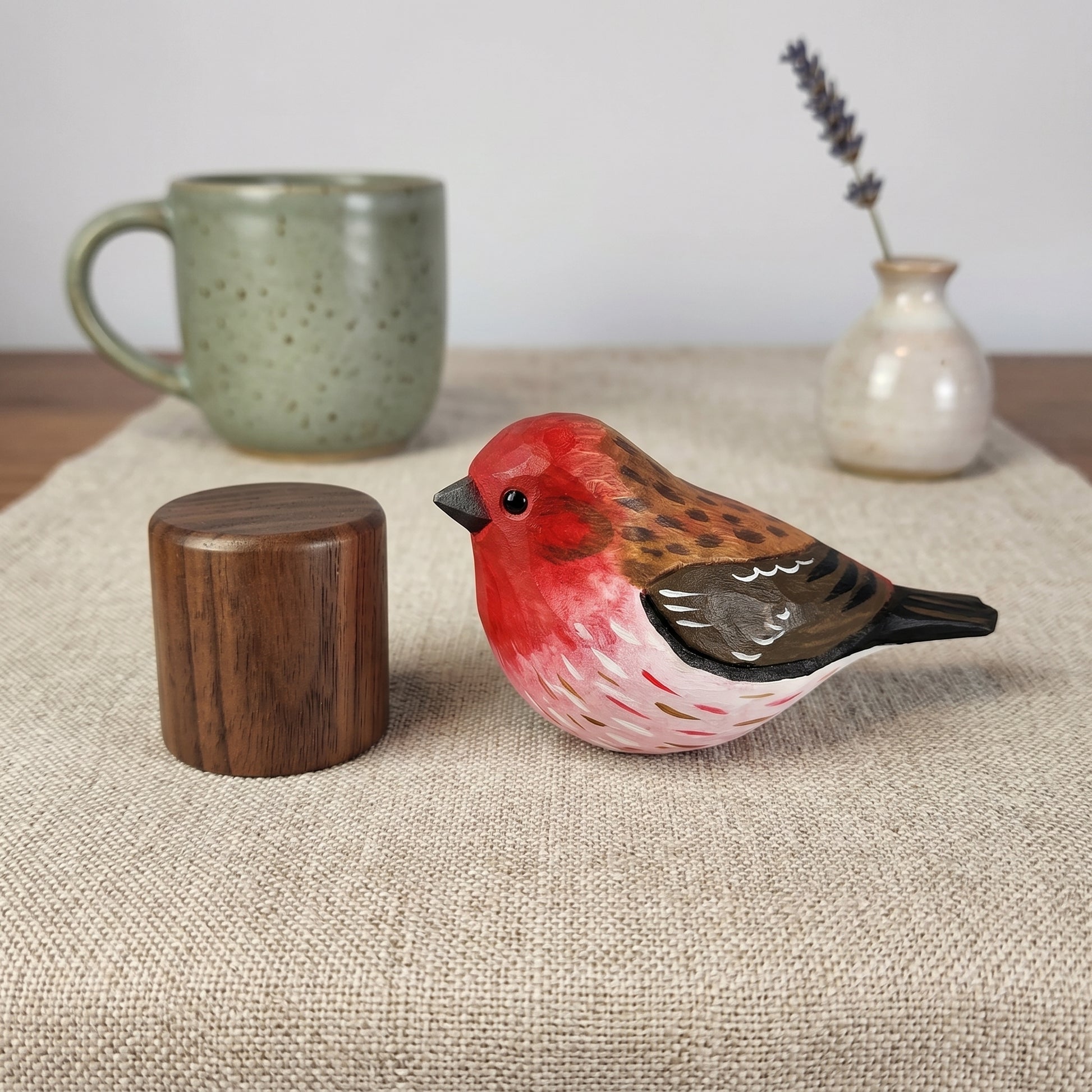 A hand-painted wooden Purple finch figurine with a red chest, brown back, and black beak, displayed on a beige linen runner next to a small wooden cylinder, with a green ceramic mug and a small vase in the background.