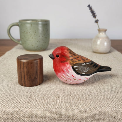 A hand-painted wooden Purple finch figurine with a red chest, brown back, and black beak, displayed on a beige linen runner next to a small wooden cylinder, with a green ceramic mug and a small vase in the background.