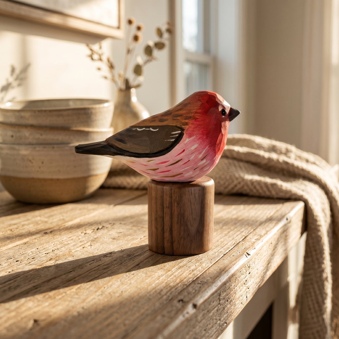 A handcrafted wooden Purple finch figurine with a pink, red, and brown painted body perched on a small cylindrical wooden base, displayed on a rustic wooden table in a warm, sunlit home setting.