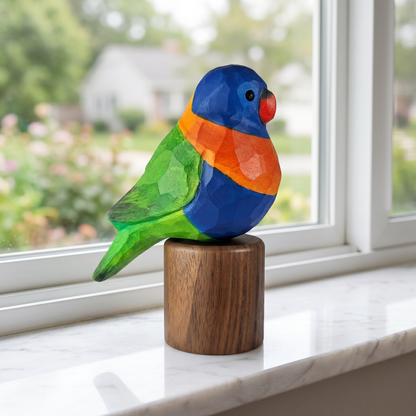 A colorful wooden Rainbow lorikeet sculpture with a blue head, green wings, orange chest, and red beak, displayed on a round wooden base on a white marble windowsill with a blurred garden view in the background.