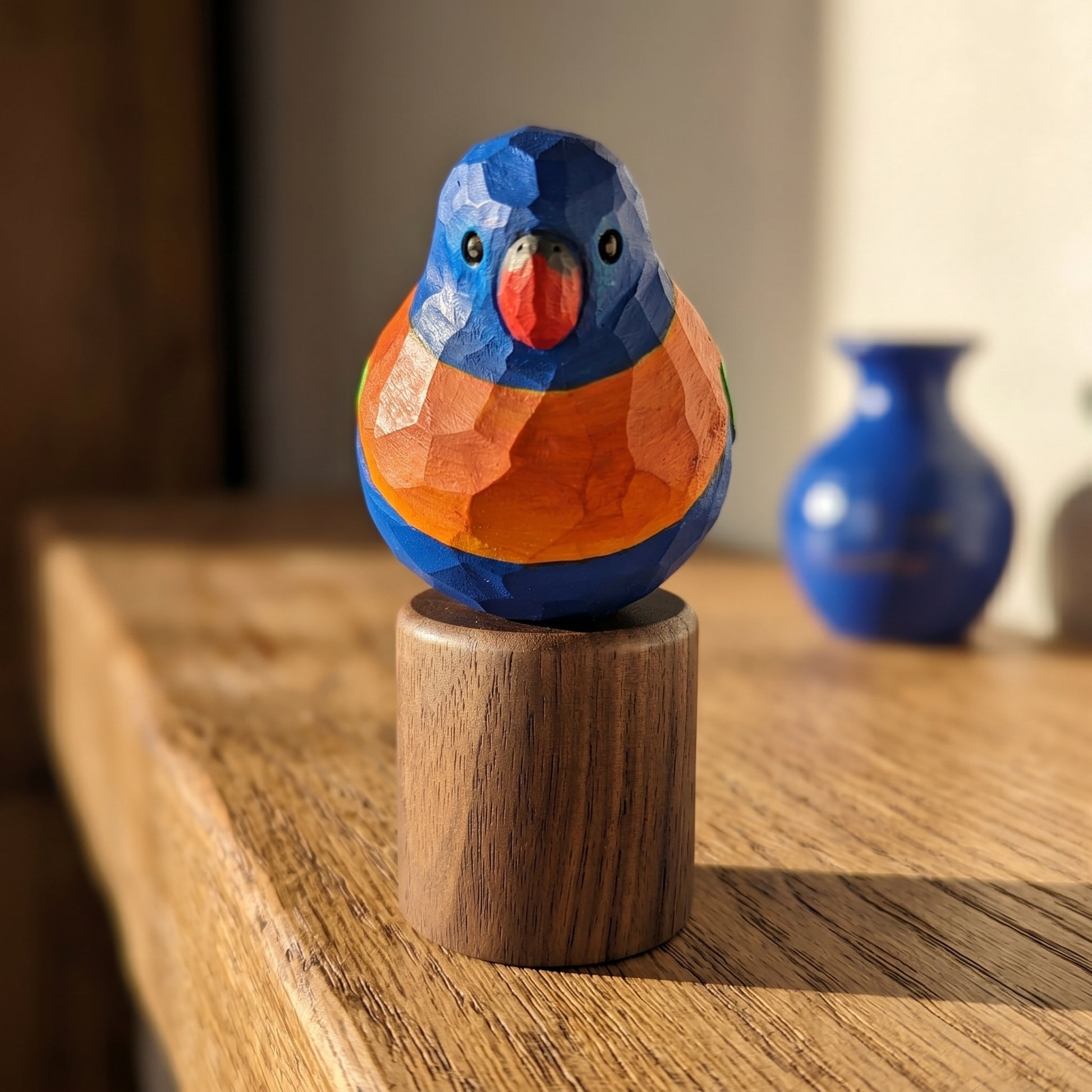 A hand-carved wooden Rainbow lorikeet figurine with a geometric, faceted finish, painted in vibrant blue, orange, and red, mounted on a small cylindrical wooden base and displayed on a wooden table.