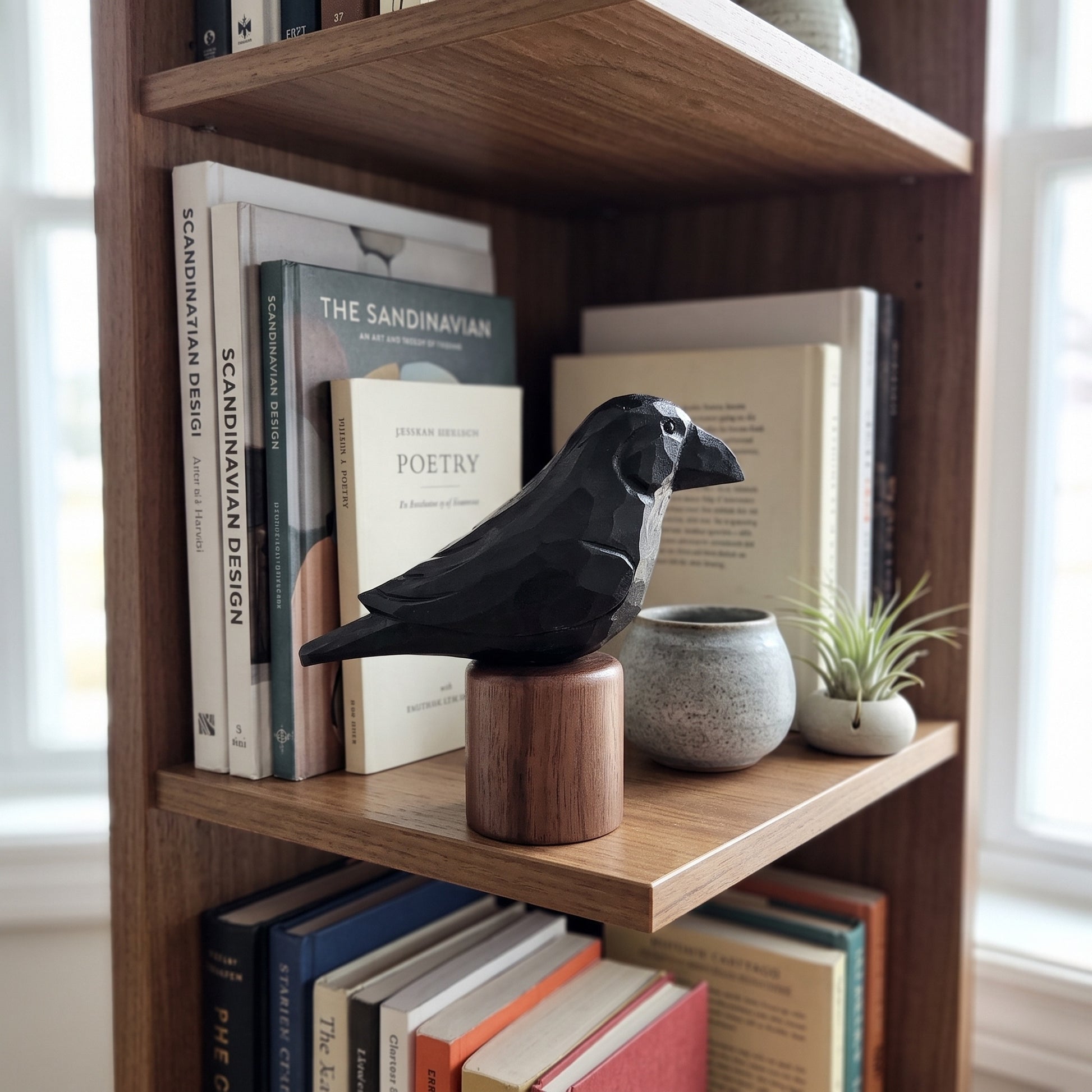 A black raven figurine mounted on a round wooden base, displayed on a wooden bookshelf alongside books, a small speckled ceramic pot, and a small potted plant.