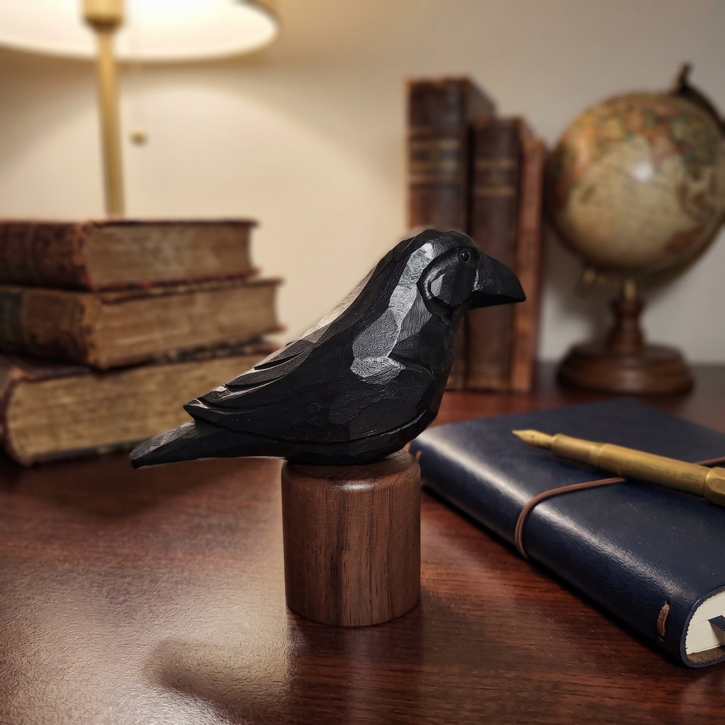 A carved black Raven figurine perched on a round wooden base, displayed on a wooden desk with vintage books, a lamp, a globe, and a navy notebook in a warm indoor setting.