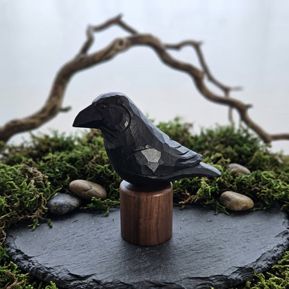 A carved black raven figurine mounted on a round wooden base, displayed on a dark stone slab surrounded by green moss and small stones, with a blurred natural branch in the background.
