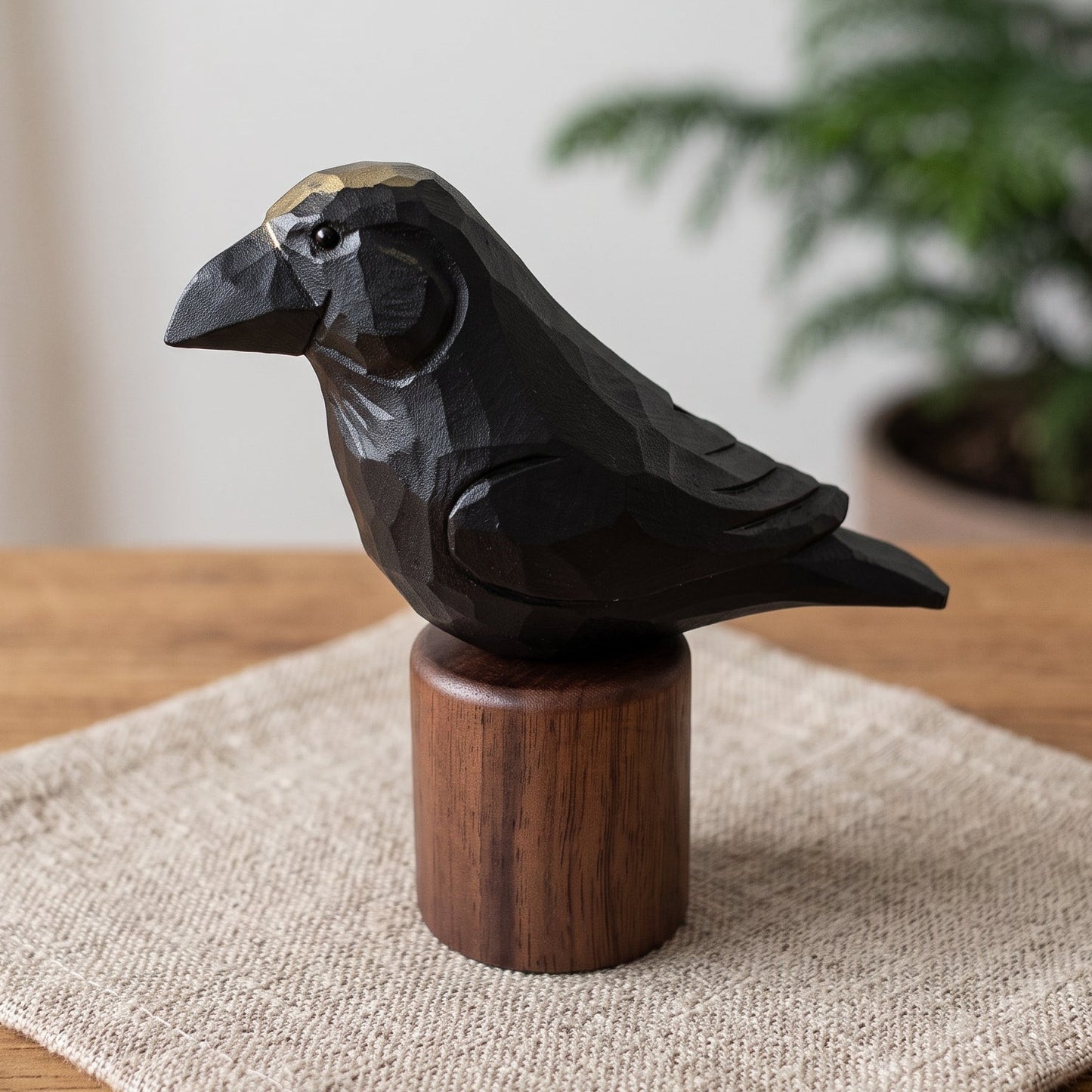 A dark carved Raven figurine with a small gold accent on its head, mounted on a cylindrical wooden base and displayed on a textured beige cloth on a wooden table with a blurred indoor plant in the background.