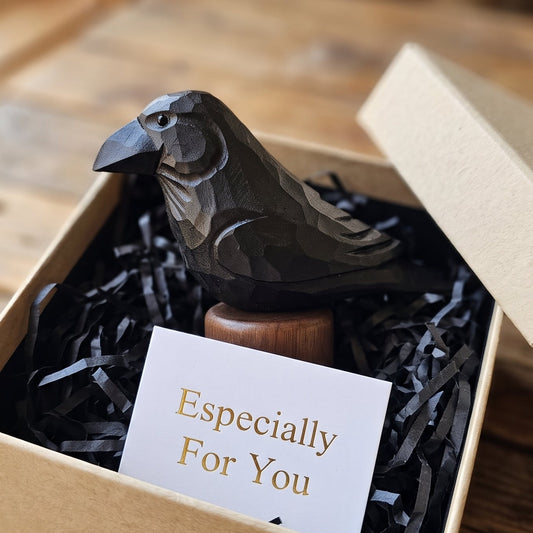 A carved wooden raven figurine displayed inside a kraft gift box with shredded black paper filler and a small card reading 'Especially For You.' The box sits on a rustic wooden table in a warm indoor setting.