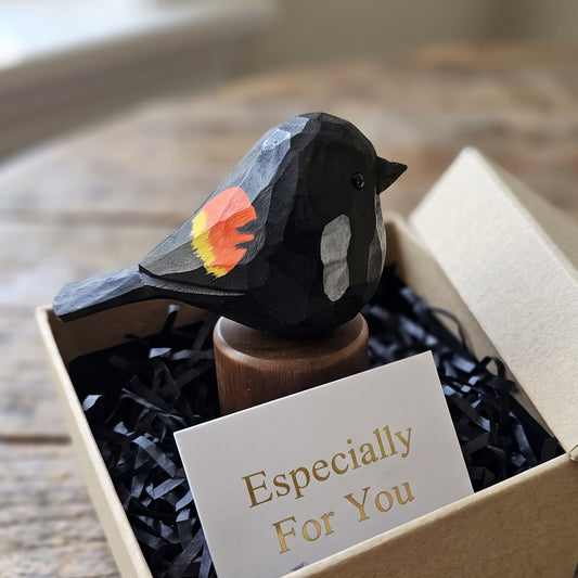 A small Red-winged blackbird figurine with orange, yellow, and white painted accents sits in a kraft gift box filled with black shredded paper and a card that reads 'Especially For You'.
