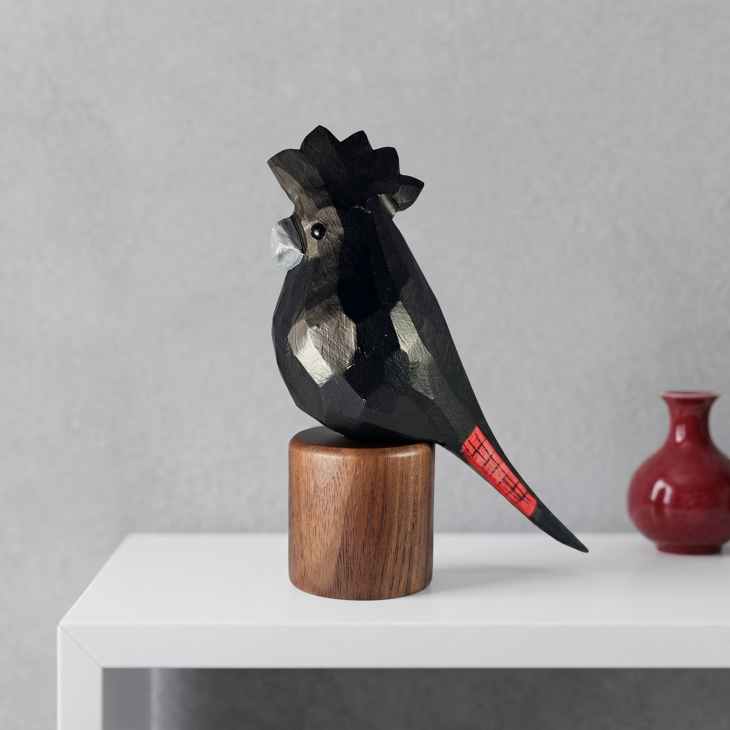 A stylized Red-tailed Black Cockatoo sculpture with geometric facets and a red tail detail, mounted on a round wooden pedestal. The sculpture is displayed on a white shelf against a gray wall, with a small red vase in the background.