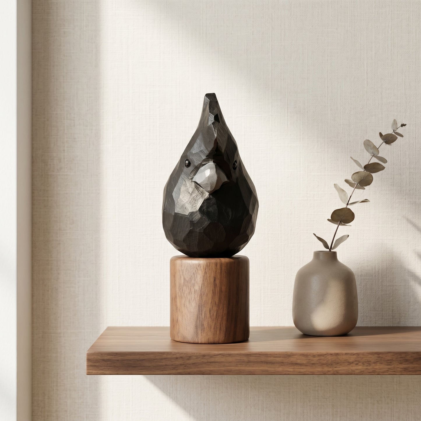 A faceted Red-tailed Black Cockatoo teardrop-shaped decorative sculpture on a cylindrical wooden base, styled on a floating wooden shelf next to a small matte beige vase with dried eucalyptus stems against a textured beige wall.