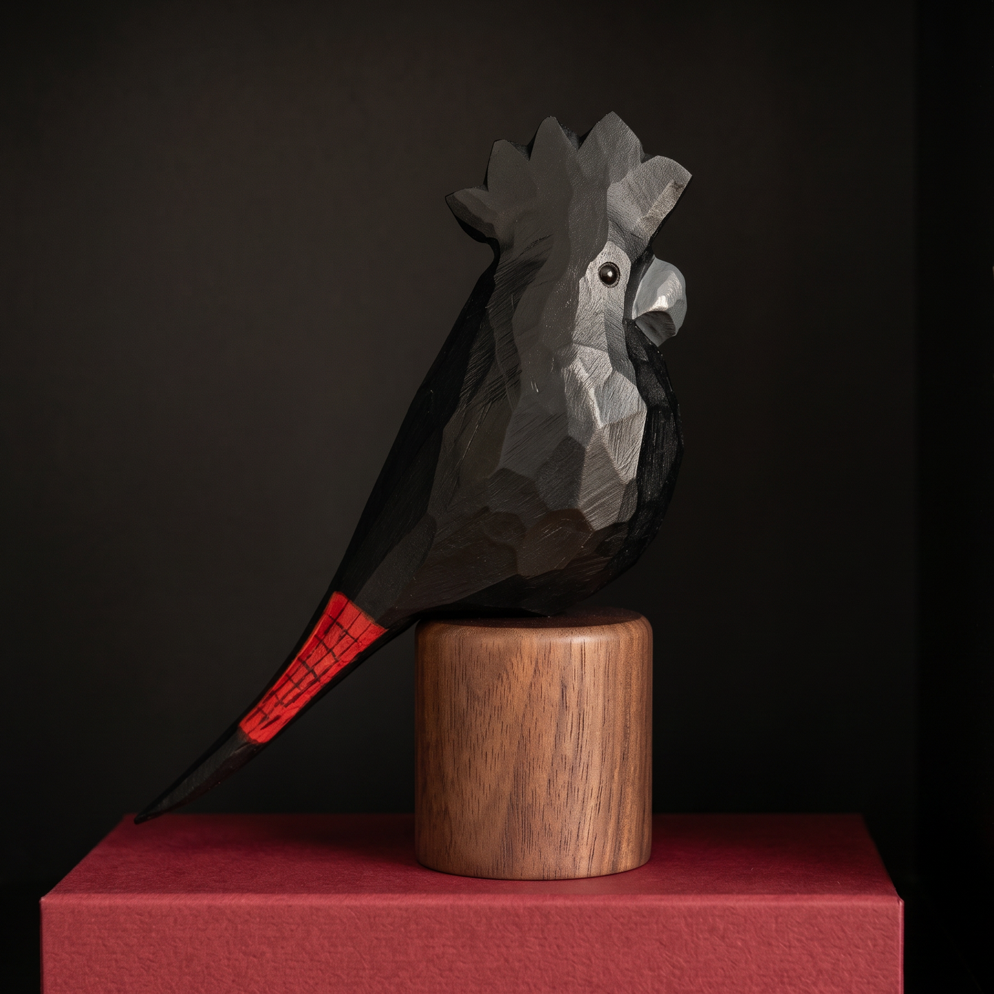 A geometric, faceted black Red-tailed Black Cockatoo sculpture with a bright red tail accent, mounted on a cylindrical wooden pedestal atop a red rectangular base, set against a dark background.