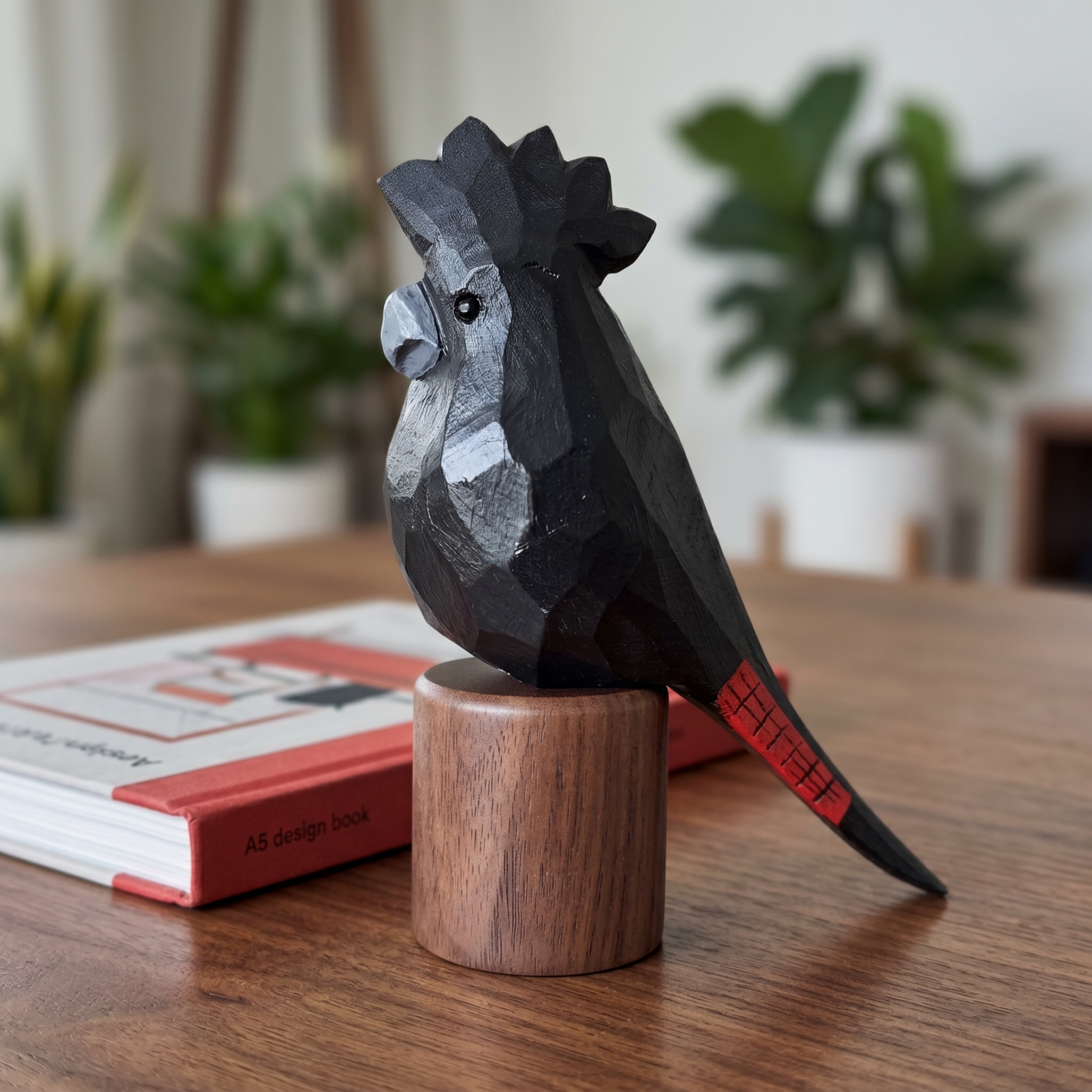 A faceted Red-tailed Black Cockatoo figurine with a red tail accent perched on a round wooden base, displayed on a wooden table next to a book with a blurred indoor plant background.