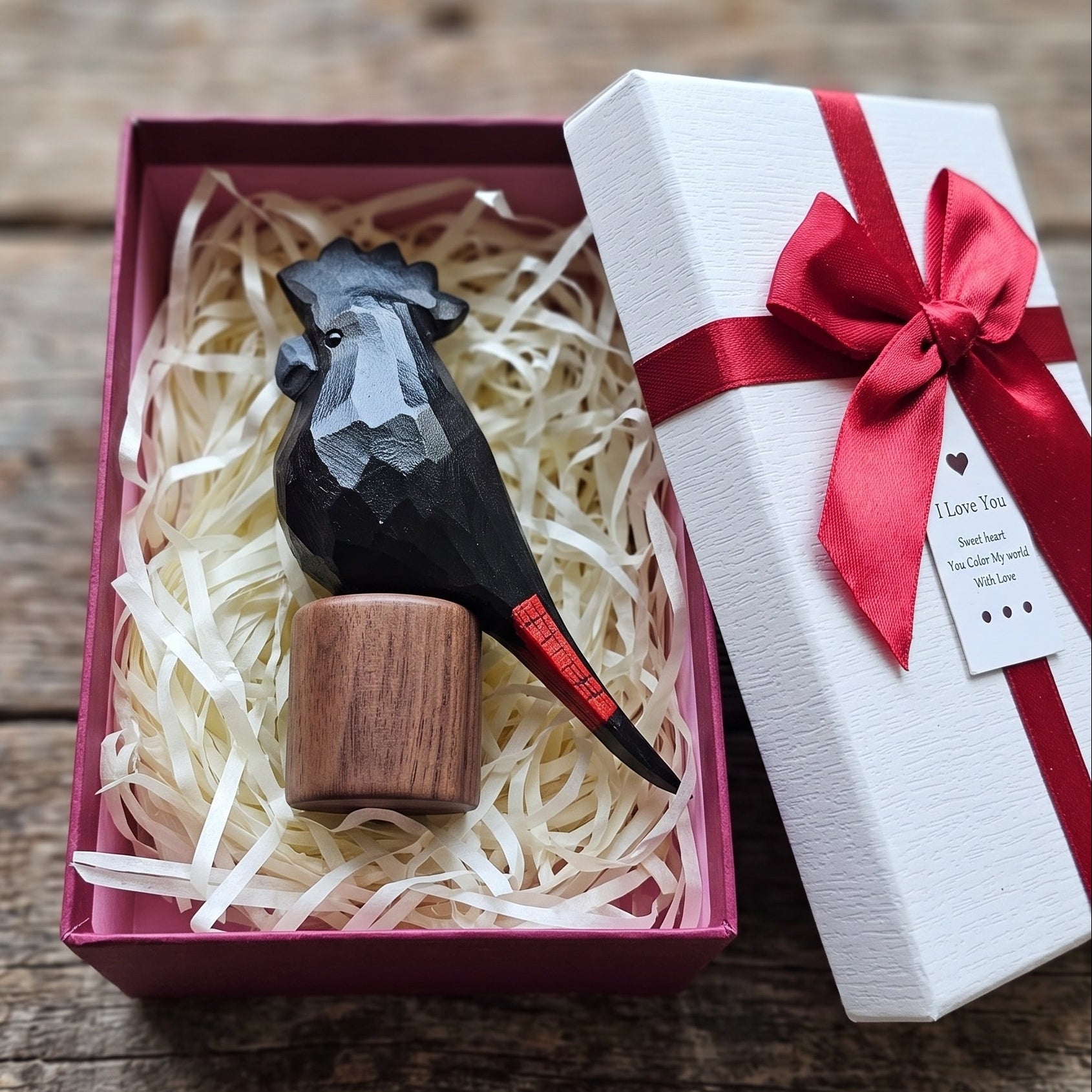 Hand-carved Red-tailed Black Cockatoo figurine on a Black Walnut base, nestled in cream paper shred inside a red gift box, with a white ribbon-tied lid and gift tag on a rustic wooden surface.