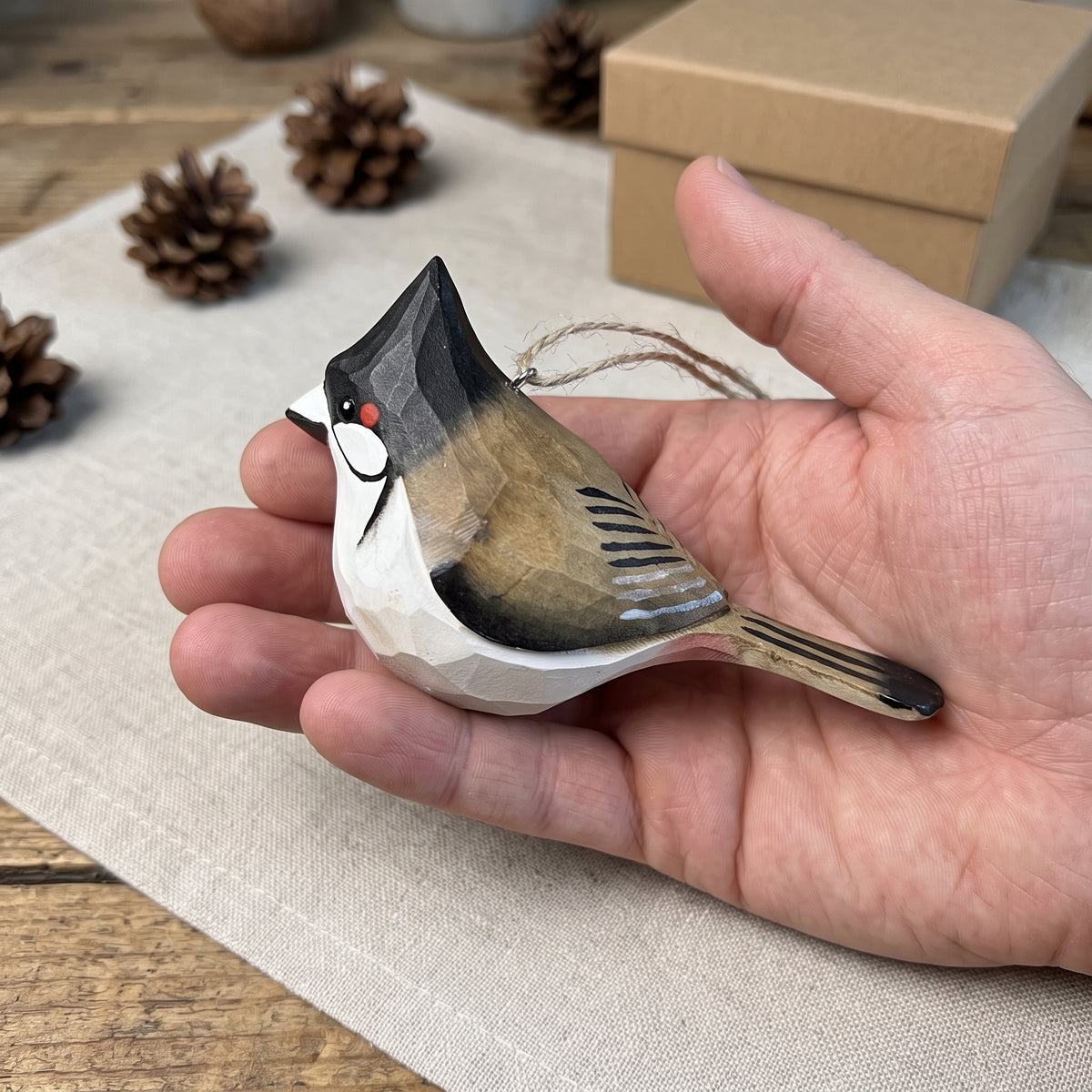 A hand holds a small carved Red-whiskered bulbul ornament with a natural wood grain finish, featuring black, white, and brown painted details. The background shows a rustic wooden table with pinecones and a kraft paper box.