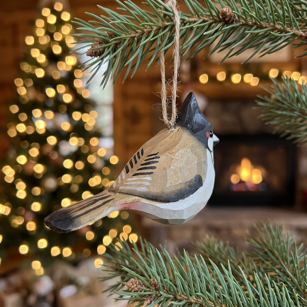 A carved wooden Red-whiskered bulbul ornament, shaped like a small bird with a black crest, white face, and brown wings, hanging from a pine branch in front of a cozy fireplace with warm lights.