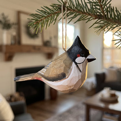 A hand-painted Red-whiskered bulbul ornament shaped like a tanager hanging from a pine tree branch in a cozy living room setting.