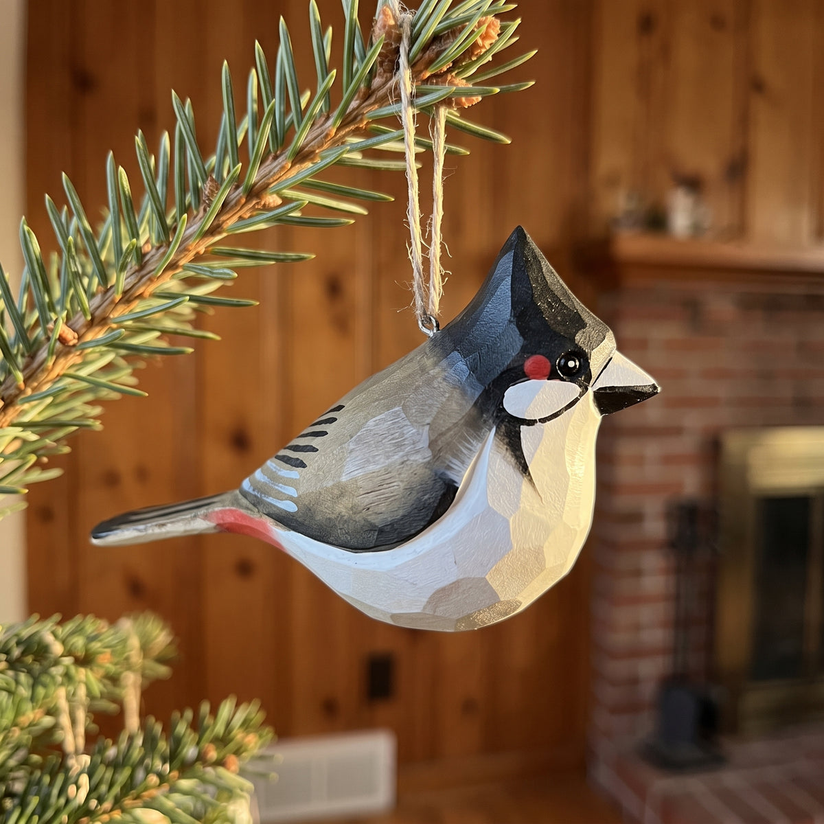 Hand-painted wooden Red-whiskered bulbul ornament shaped like a blue tit, hanging from a pine branch in a cozy indoor setting with a wood-paneled wall and fireplace in the background.