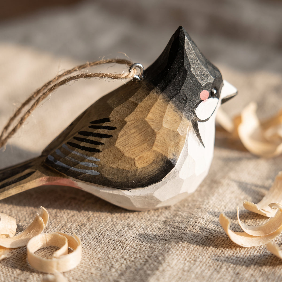 A handcrafted wooden Red-whiskered bulbul ornament with geometric facets, painted in black, brown, and white with a small pink accent, hanging from a twine string on a burlap surface with wood shavings.