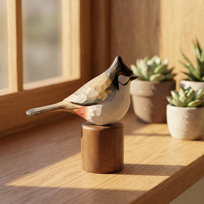 A geometric Red-whiskered bulbul figurine with a black, white, and gray faceted pattern and a small red eye, standing on a round wooden base on a sunlit wooden windowsill with potted succulents in the background.