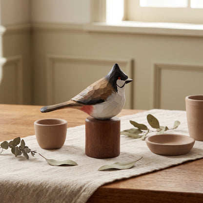 A carved wooden Red-whiskered bulbul figurine with a dark crest and red chest sits on a cylindrical wooden pedestal on a dining table. The scene is styled with matching terracotta bowls, a linen runner, and scattered eucalyptus leaves in a softly lit room.
