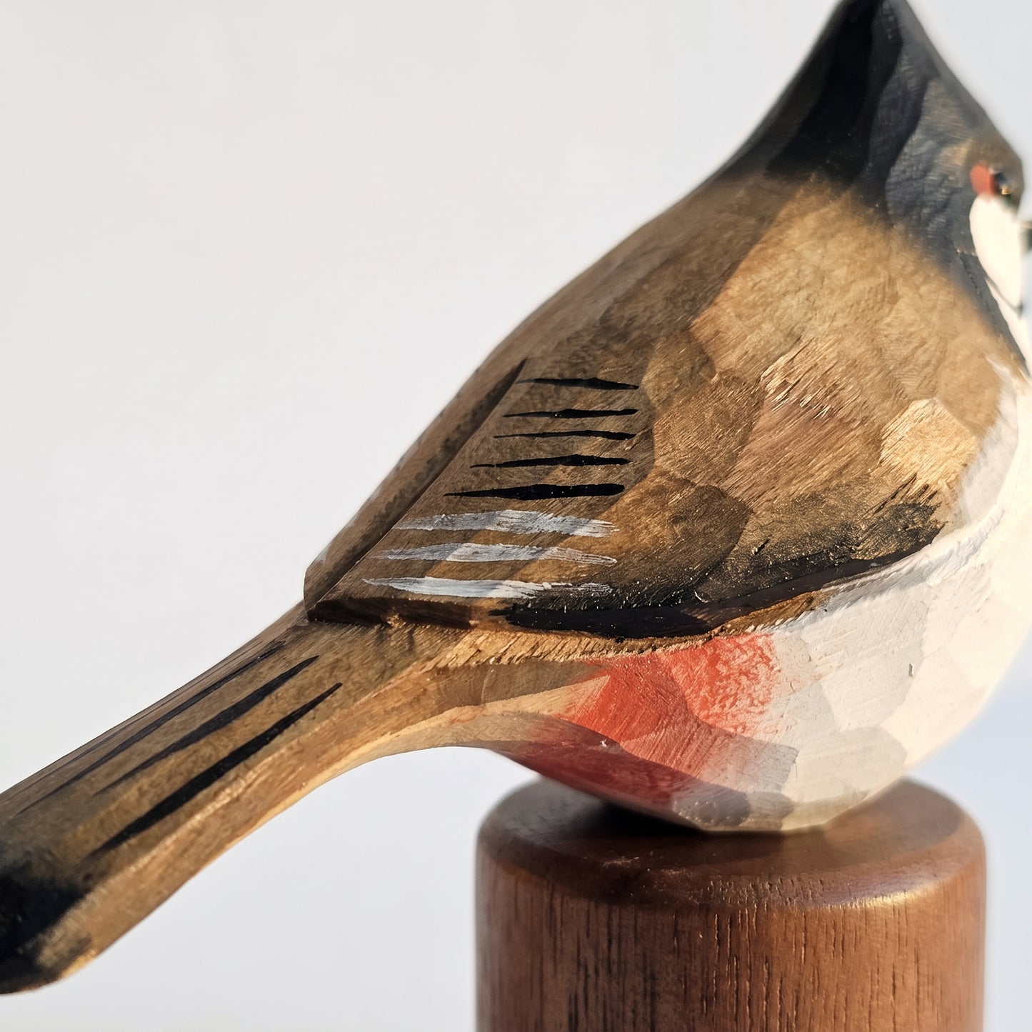 Close-up of a carved wooden Red-whiskered bulbul figurine perched on a round wooden base, showing natural wood grain and hand-carved details against a plain light background.