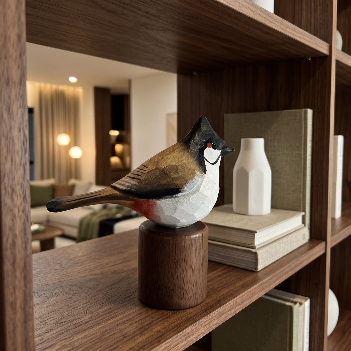 A geometric Red-whiskered bulbul sculpture with a faceted white, brown, and black design sits on a cylindrical wooden base, displayed on a dark wood bookshelf next to books and a white vase.