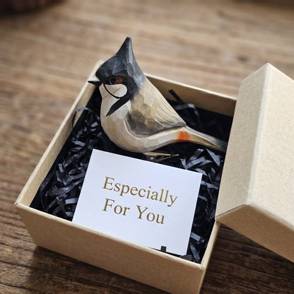 A hand-carved wooden Red-Whiskered Bulbul ornament displayed inside an open kraft gift box filled with black shredded paper, featuring a tall pointed black crest, crimson whisker patch, white cheek, dark brown wing, red undertail accent, and a white card reading "Especially For You" placed in the foreground.