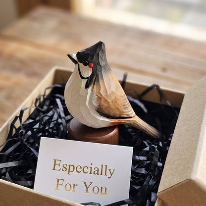 A small carved Red-whiskered bulbul figurine sits in a kraft gift box filled with black paper shred, with a card reading 'Especially For You' in front. The box is placed on a wooden table in a softly lit indoor setting.