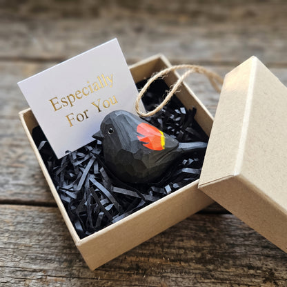 Hand-carved wooden Red-Winged Blackbird hanging ornament packaged in an open kraft gift box with shredded paper fill and an "Especially For You" card.