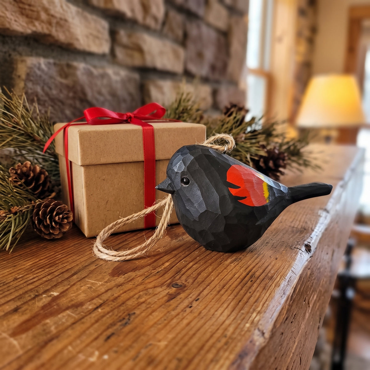 A black faceted Red-Winged Blackbird  Christmas ornament with a red and yellow wing accent and a jute hanging loop, displayed on a rustic wooden mantel next to a kraft gift box with a red ribbon, pinecones, and evergreen branches in front of a stone fireplace.