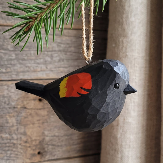 A handcrafted wooden Red-winged blackbird ornament in black with a red and yellow accent on its wing, hanging from a pine branch with twine against a rustic wood and burlap background.
