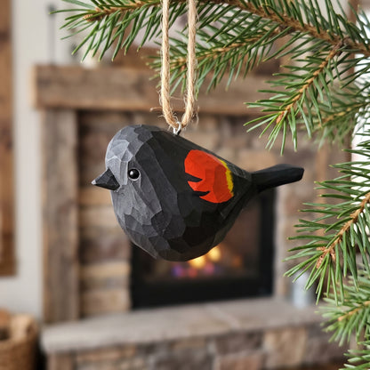 Hand-carved wooden Red-Winged Blackbird ornament hanging from a pine branch by a twine loop in a rustic indoor setting.