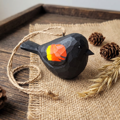 A black faceted Hand-carved wooden Red-Winged Blackbird hanging ornament held in a hand wearing a cozy knit sweater. ornament with a colorful orange, yellow, and red geometric tail, hanging from a natural twine string on a burlap fabric over a wooden tray, styled with pinecones and dried wheat.