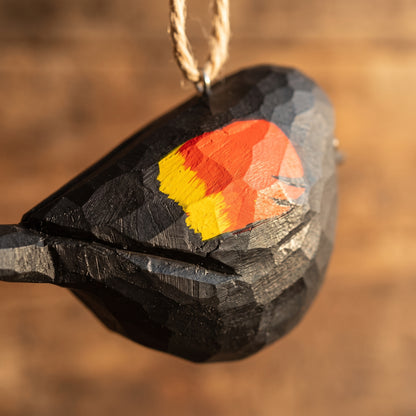 Extreme close-up of a hand-carved wooden Red-Winged Blackbird ornament highlighting the faceted chiseled wood grain and vibrant hand-painted red, orange, and yellow wing patch.