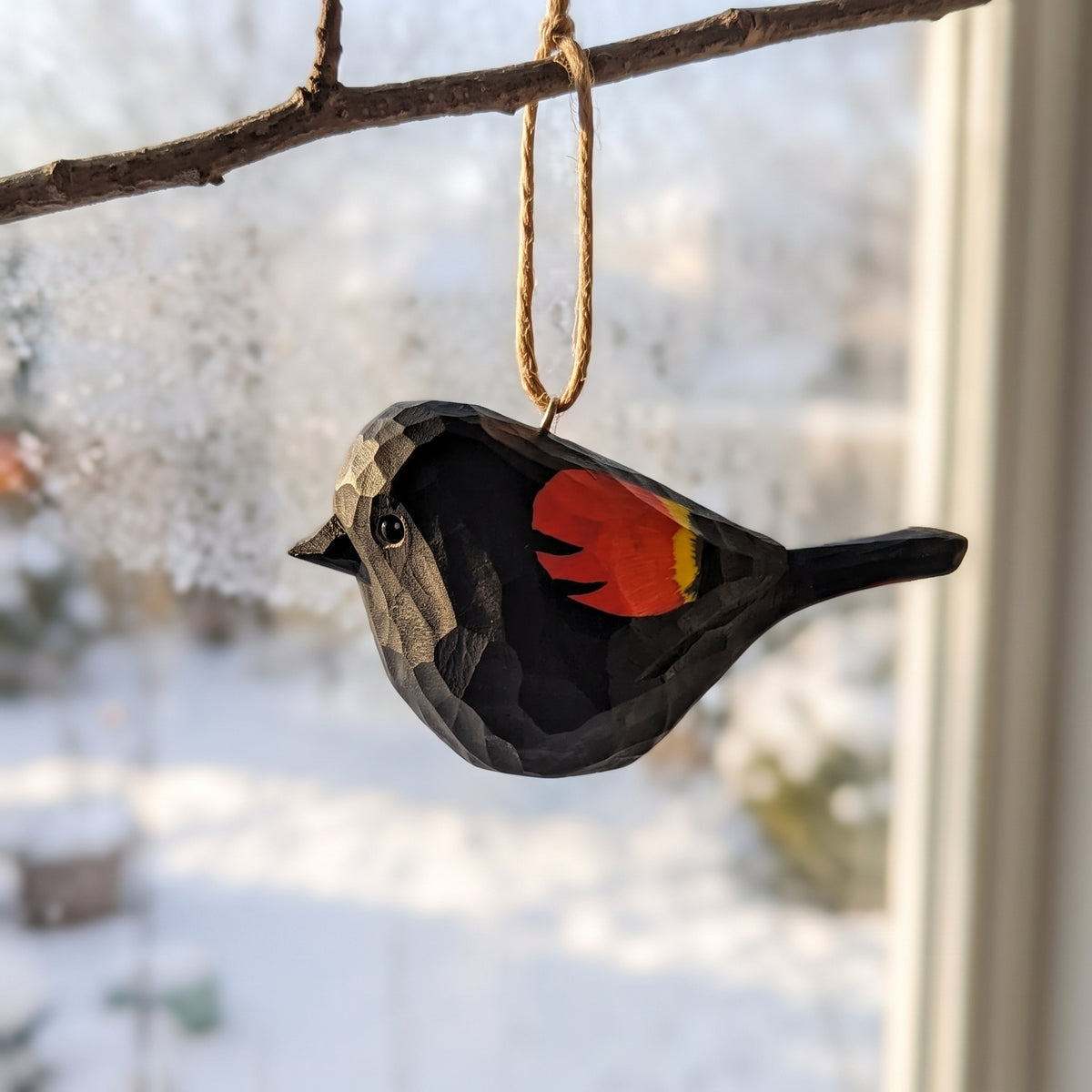 A carved wooden Red-Winged Blackbird  ornament, resembling a chickadee with a black body, gray beak and wing, and a bright red and yellow chest patch, hanging from a natural twine loop on a tree branch in front of a snowy window.