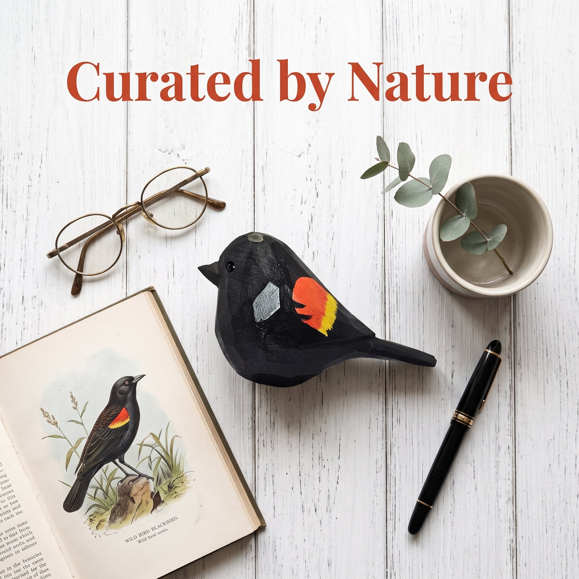 A styled flat-lay featuring a Red-winged blackbird-shaped object with red and orange feather details, arranged on a white wood background with an open book, eyeglasses, a small cup with eucalyptus, and a black pen. The text 'Curated by Nature' appears at the top.