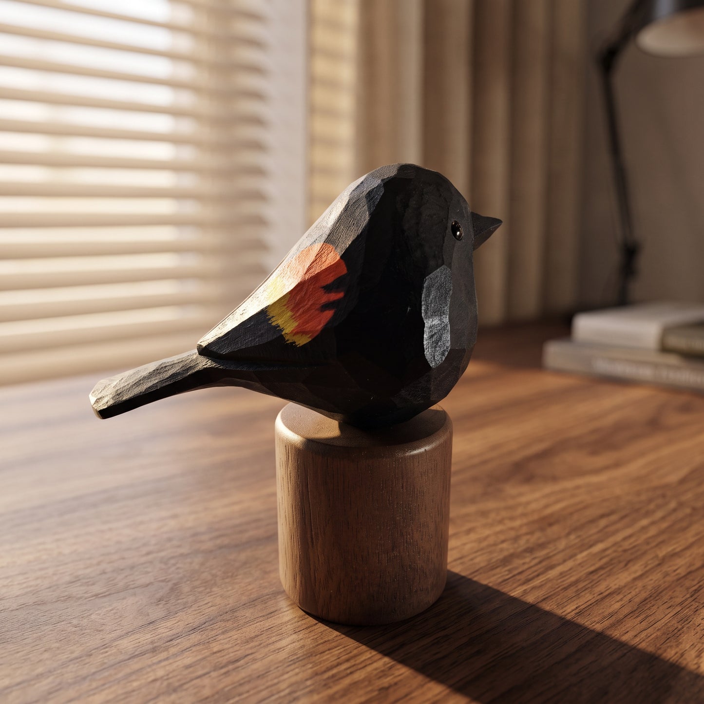 A hand-carved wooden Red-winged blackbird figurine with a black body, gray beak and cheek, and a red and yellow wing patch, mounted on a cylindrical wooden base on a wooden table.