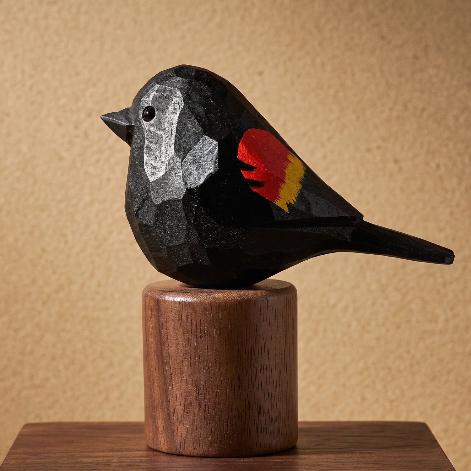 A carved Red-winged blackbird sculpture with a black body, white face, and red and yellow wing detail, mounted on a cylindrical wooden pedestal against a warm beige textured background.