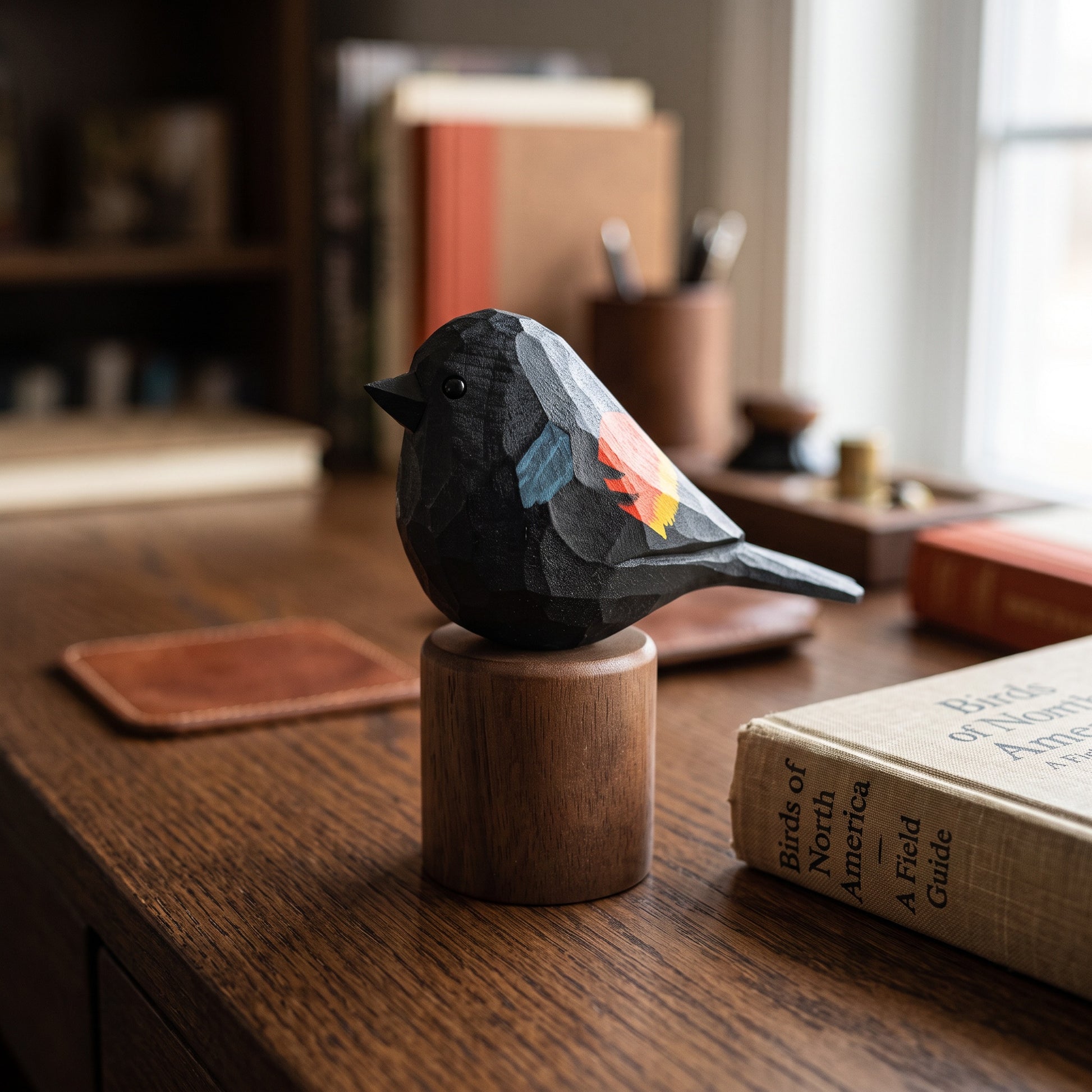 A small carved Red-winged blackbird figurine with dark charcoal and gray tones, featuring painted accents of orange, yellow, and light blue on its wing and back, displayed on a round wooden pedestal on a wooden desk.