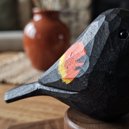 A close-up of a hand-carved Red-winged blackbird sculpture with a painted red and yellow chest accent, resting on a wooden surface with a blurred terracotta vase in the background.