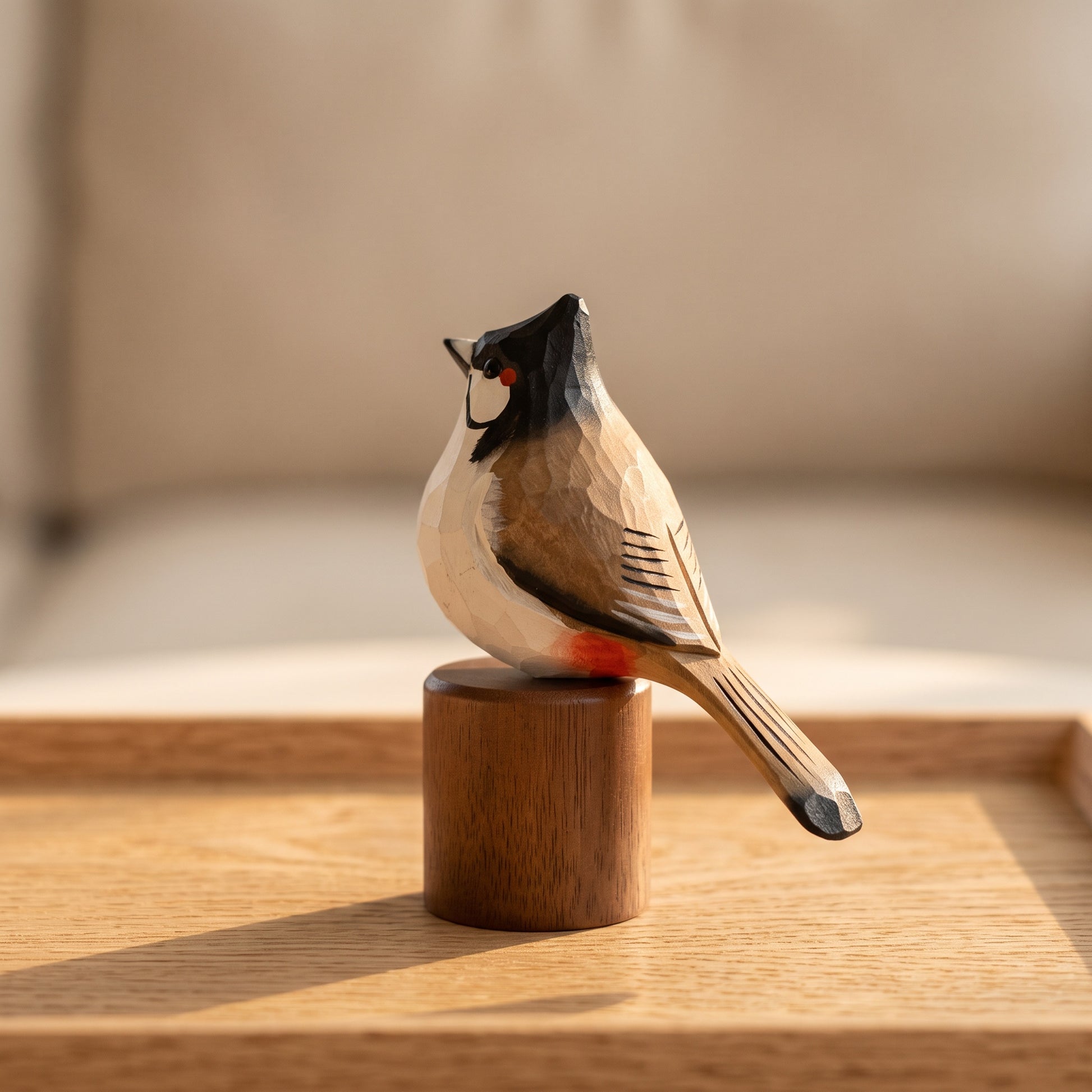 A hand-carved wooden Red-whiskered bulbul figurine with a dark crest, cream belly, and reddish-orange underbelly, perched on a small cylindrical wooden base on a light wood surface.