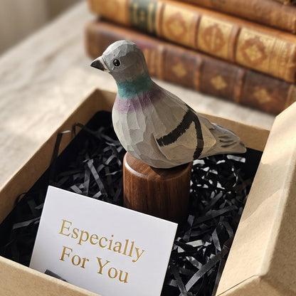 A small carved Rock dove figurine on a wooden stand presented in a kraft gift box with black shredded paper and a card that reads 'Especially For You.' Stacked vintage books sit blurred in the background on a table.