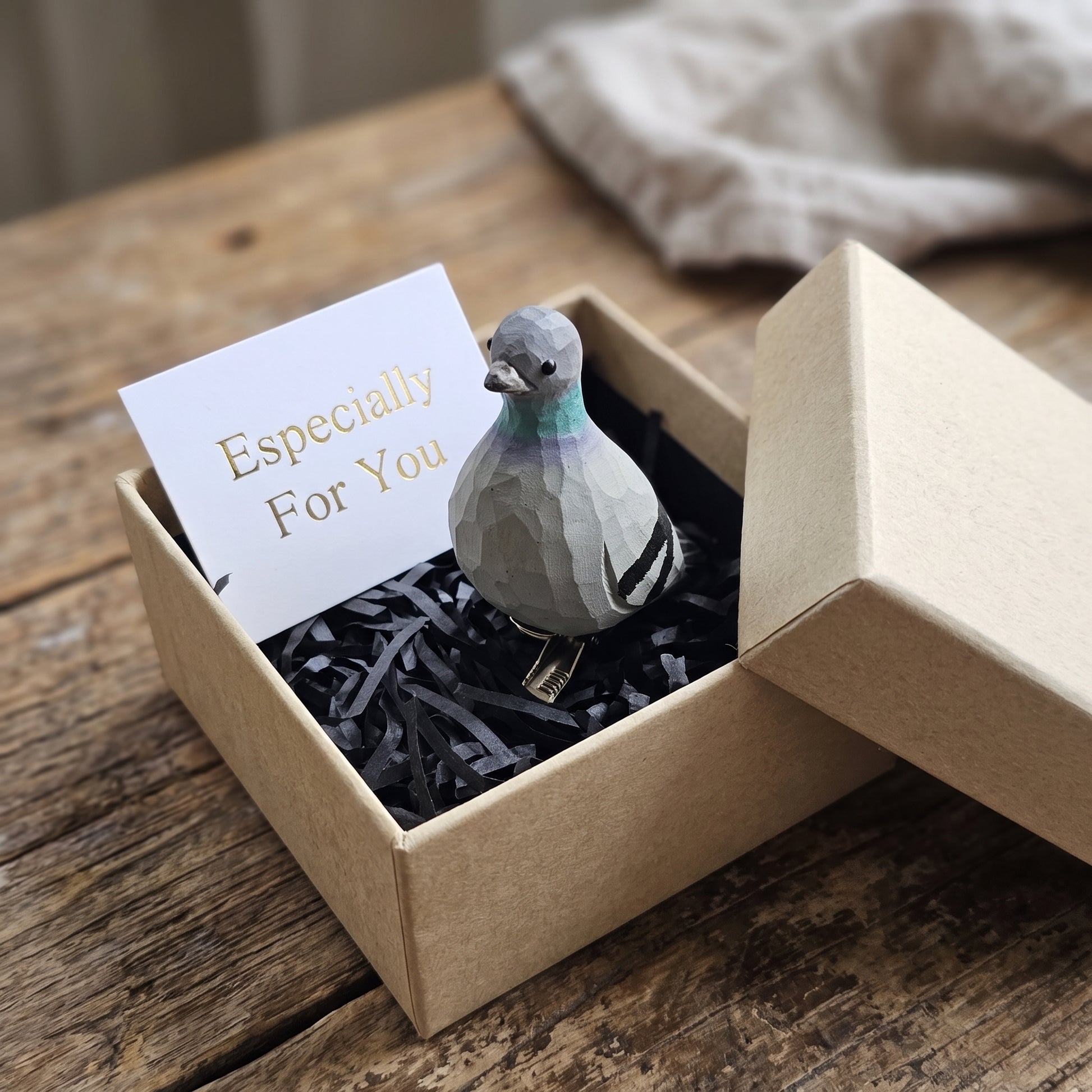 Hand-carved wooden Rock Dove (pigeon) bird ornament perched in black shredded paper inside an open kraft gift box, next to a white card reading 'Especially For You'.