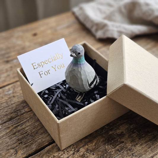 Hand-carved wooden Rock Dove (pigeon) bird ornament perched in black shredded paper inside an open kraft gift box, next to a white card reading 'Especially For You'.