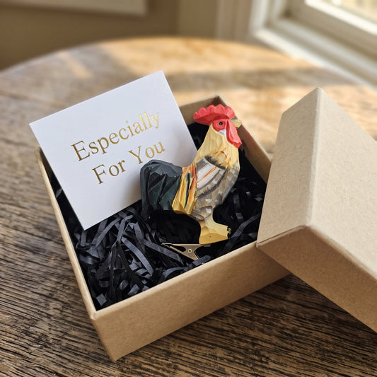 A hand-carved wooden rooster clip-on ornament resting in an open kraft gift box filled with black shredded paper, featuring a pale yellow neck, grey and white wing feathers, a dark tail, a silver metal clip underneath, and a white card reading "Especially For You" placed beside it.