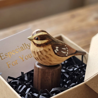 A small carved Rose-breasted Grosbeak figurine perched on a wooden stand, presented inside a kraft gift box with black shredded paper and a card that reads 'Especially For You', set on a rustic wooden table.