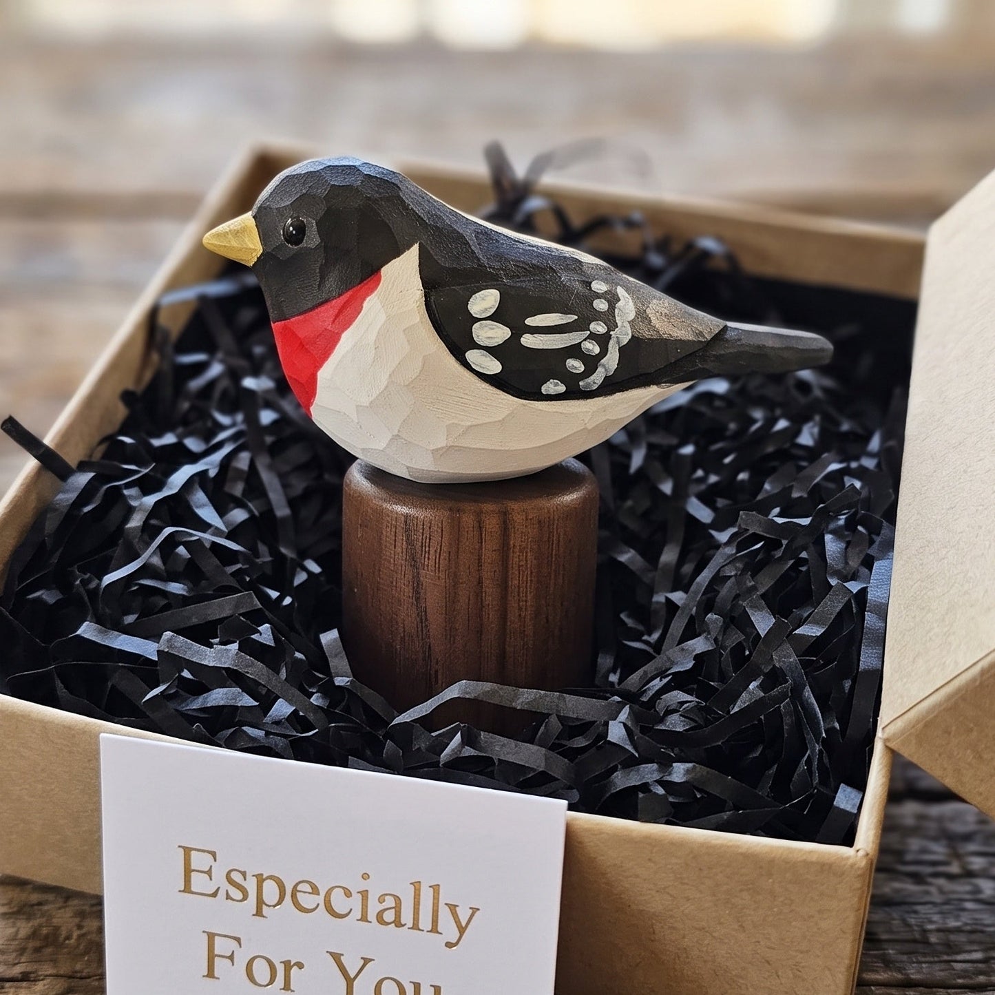 A handcrafted wooden Rose-breasted Grosbeak figurine with a red chest, black and white patterned wings, and a yellow beak, displayed in a brown kraft gift box filled with black shredded paper. A small card reading 'Especially For You' is placed in front.
