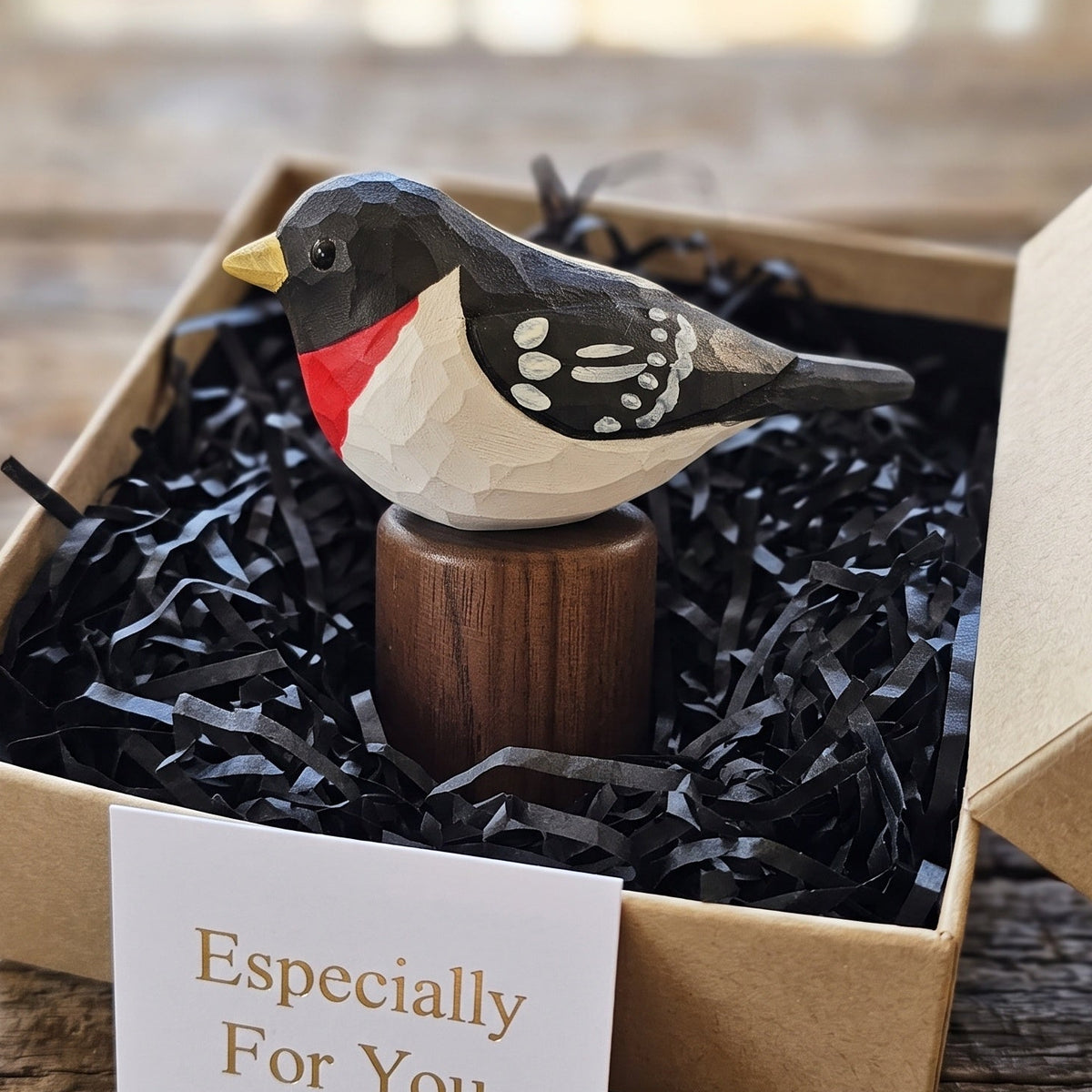 A handcrafted wooden Rose-breasted Grosbeak figurine with a red chest, black and white patterned wings, and a yellow beak, displayed in a brown kraft gift box filled with black shredded paper. A small card reading 'Especially For You' is placed in front.