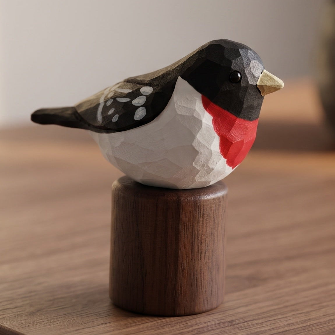 A geometric, faceted wooden Rose-breasted Grosbeak figurine with a black, white, and red painted design, perched on a cylindrical dark brown wooden base, set on a wooden tabletop.